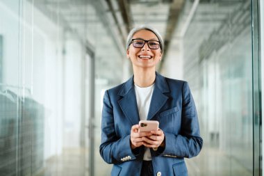 Mutlu, beyaz saçlı, olgun iş kadını gülüyor ve cep telefonunu içeride kullanıyor.