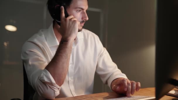 一个自信英俊的男人打电话给某人 看着办公室里的电脑屏幕的侧面视图 — 图库视频影像