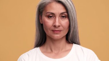 Kendinden emin, olgun bir Asyalı kadın bej rengi stüdyoda kameraya gözlerini açıyor.