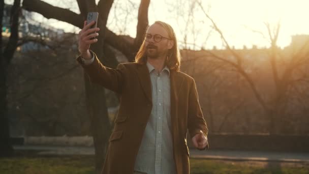 コートを着た笑顔の長髪の男がセルフィーを作り 日没の背景に対して様々なジェスチャーを示しています — ストック動画