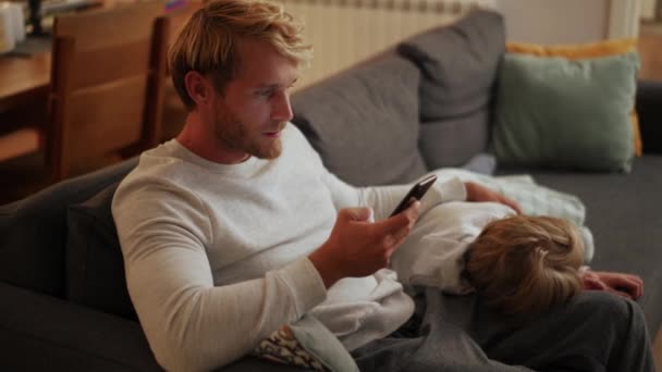 息子がソファの上で眠っている間に電話で集中的な父のテキストメッセージの側面図 — ストック動画