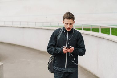 Stadyumda yürüyen, spor çantası taşıyan, cep telefonu kullanan kendinden emin genç bir adam.