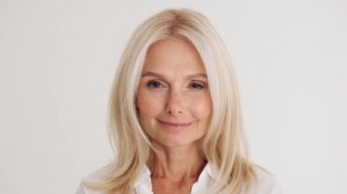 Beyaz stüdyoda gülümseyen olgun kadının kameraya gözlerini açtığı yakın çekim.