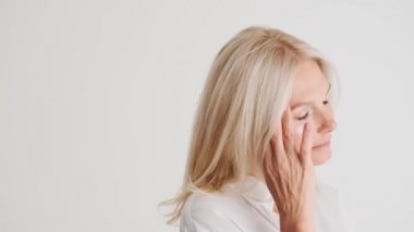 Beyaz stüdyoda yüzüne dokunan konsantre olmuş olgun kadının yan görüntüsü.