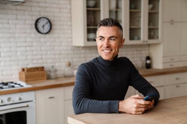 Ev mutfağında otururken cep telefonu kullanan yakışıklı adam.