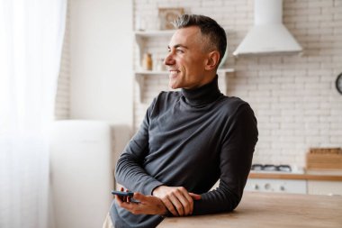 Ev mutfağında masaya yaslanırken cep telefonu kullanan yakışıklı adam.