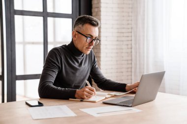 Masada otururken bilgisayar ve kağıtlarla uğraşan yakışıklı bir adam.