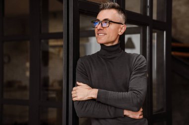 Gözlüklü yakışıklı adam evde duvara yaslanırken kameraya gülümsüyor.