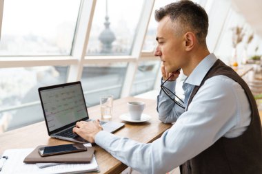 Kendine güvenen, şık giyimli yakışıklı iş adamı kafede otururken dizüstü bilgisayarla çalışıyor.