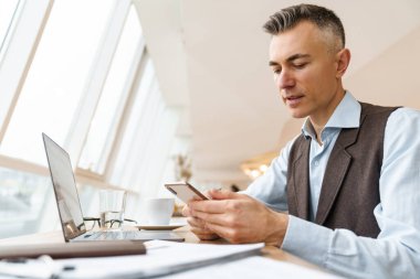 Kendine güvenen yakışıklı iş adamı laptopta çalışırken kafede oturuyor, cep telefonu kullanıyor, kahve içiyor.