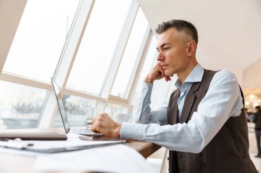 Kendine güvenen, şık giyimli yakışıklı iş adamı kafeteryada otururken dizüstü bilgisayarla çalışıyor.