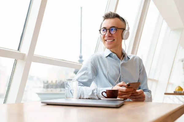 Handsome smiling man in headphones listening to music, using a smartphone, drinking coffee while resting in cafe