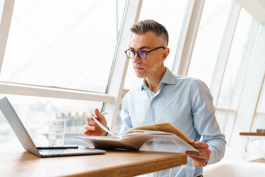 Confident handsome businessman in smart clothes working on laptop computer while sitting in the cafe indoors, looking through documents
