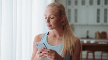 Gülümseyen sarışın kadın evde telefonla mesaj atıyor.