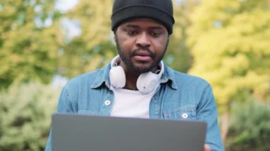 Kulaklıklı konsantre Afrikalı adam parkta dizüstü bilgisayarından bir şey okuyor.