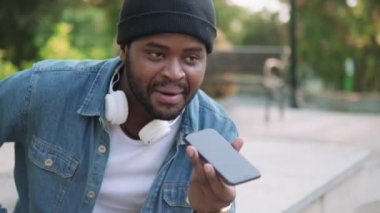 Kulaklıklı mutlu Afrikalı adamın yakın plan görüntüsü beton bir bankta otururken telefonla sesli mesaj kaydediyor.