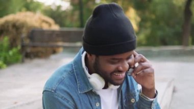 Kulaklıklı gülen Afrikalı adamın beton bir bankta otururken telefonla konuşmasına yakından bakın.