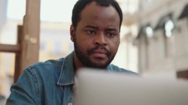 Laptop üzerinde çalışan ve açık kafede yazan dalgın Afrikalı adamın yakın çekim görüntüsü.