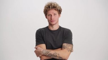 Beyaz stüdyoda kendine güvenen kıvırcık saçlı, tişörtlü bir adam kameraya bakıyor.