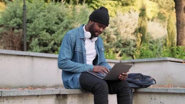 Hombre Africano Positivo Auriculares Mensajes Texto Por Ordenador Portátil Banco — Vídeos de Stock