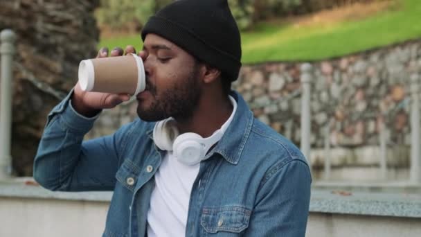 Konzentrierter Afrikaner Mit Kopfhörern Der Kaffee Auf Einer Betonbank Trinkt — Stockvideo