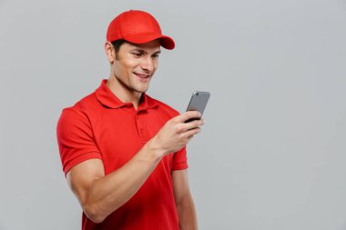 Beyaz duvardan izole edilmiş cep telefonu kullanırken gülümseyen üniformalı genç teslimatçı.