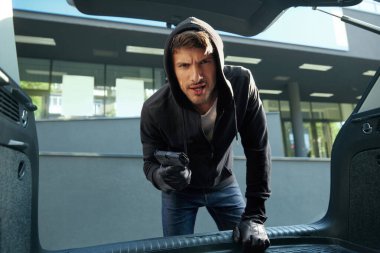 Kızgın Avrupalı soyguncu arabanın bagajına bakıyor. İnsan kurbanla birlikte bagajın görüntüsü. Erkek sakallı haydut siyah kapüşonlu eldiven takıyor ve tabanca tutuyor. Adam kaçırma kavramı. Şehir gündüz vakti