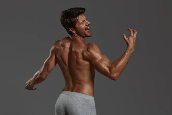 Young beautiful screaming european athletic man. Back view of bearded man with dark hair and naked sportive body. Isolated on dark gray background. Studio shoot. Copy space