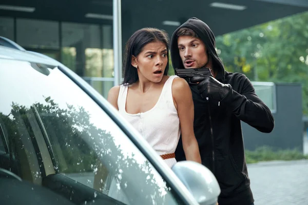 Angry robber threatening with pistol to frightened girl. European man and young brunette woman stand near automobile. Male bandit wear hoodie. Concept of robbery and kidnapping. City daytime