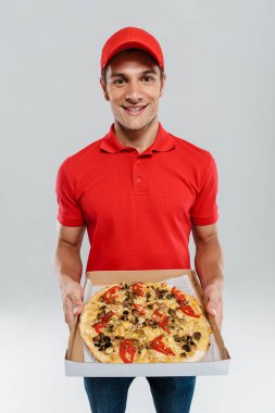 Beyaz duvarda izole edilmiş pizzayı gösterirken üniformalı genç teslimatçı gülümsüyor.