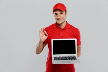 Genç teslimatçı, beyaz duvarın üzerinde dizüstü bilgisayar gösterirken el kol hareketi yapıyor.