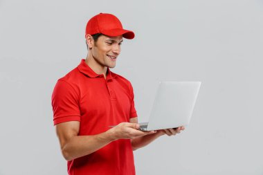 Beyaz duvarın üzerinde dizüstü bilgisayarla poz verirken gülümseyen üniformalı genç teslimatçı.