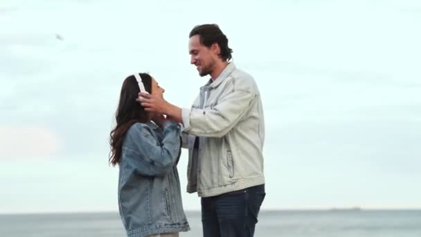 一个戴着耳机的帅哥给他的女朋友 女朋友开始在海边跳舞 — 图库视频影像