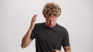 Beyaz stüdyoda telefonla mesajlaşan sinirli kıvırcık saçlı adam.