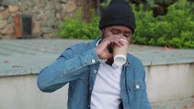 Kulaklıklı kendinden emin Afrikalı adam beton bir bankta kahve içiyor.