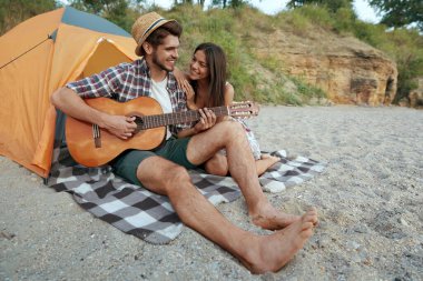 Mutlu adam, güzel gülümseyen kıza gitarla çalar. Genç Avrupalı aşık çift kumsalda dinleniyor. Romantik ilişki ve birlikte vakit geçirme kavramı. Yaz tatilinde kamp yapmak