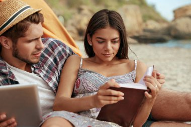 Genç Avrupalı çift kumsalda dinleniyor. Ciddi adamın elinde dijital tablet var. Odaklanmış kız kitap okuyor. Romantik ilişki ve birlikte vakit geçirme kavramı. Yaz mevsiminde doğada tatil