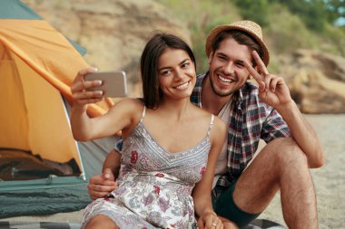 Mutlu genç çift sahilde cep telefonuyla selfie çekiyor. Adam zafer işareti gösteriyor. Romantik ilişki ve birlikte vakit geçirme kavramı. Yaz zamanı doğada kamp yapmak.