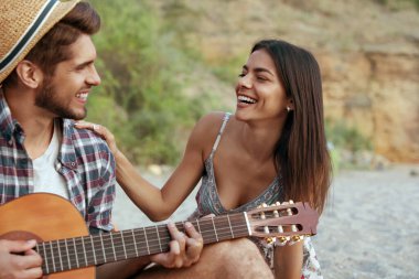 Mutlu adam, güzel ve mutlu kıza gitarla çalar. Genç Avrupalı çift kumsalda dinleniyor. Romantik ilişki ve birlikte vakit geçirme kavramı. Doğada tatil. Yaz zamanı.