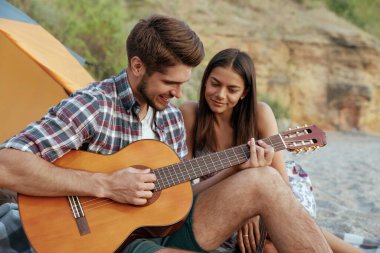 Mutlu adam, güzel gülümseyen kıza gitarla çalar. Genç Avrupalı çift kumsalda dinleniyor. Romantik ilişki ve birlikte vakit geçirme kavramı. Doğada kamp yapmak. Yaz zamanı.