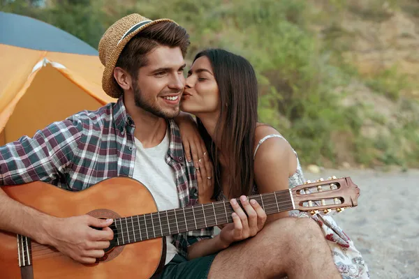 喜欢的家伙用吉他弹奏正在吻他的漂亮姑娘 一对年轻的欧洲夫妇在沙滩上休息 浪漫关系的概念和享受在一起的时光 暑期野营度假 — 图库照片