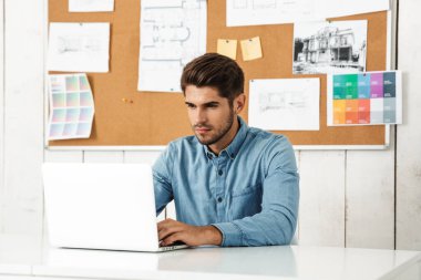 Genç esmer adam ofisteki masada otururken dizüstü bilgisayarla çalışıyor.