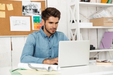 Genç esmer adam ofisteki masada otururken dizüstü bilgisayarla çalışıyor.
