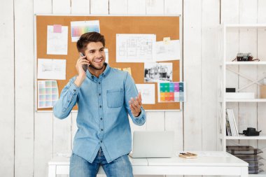 Genç esmer adam ofisteki masada otururken cep telefonuyla konuşuyor.