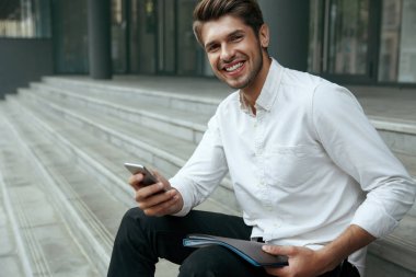 Avrupalı işadamı merdivenlerde oturuyor ve cep telefonu kullanıyor. Elinde belgeler olan genç bir adam. Şehirdeki modern ofis binasının arka planı. Gülümseyen sakallı adam beyaz gömlek ve kot pantolon giyiyor. Gündüz