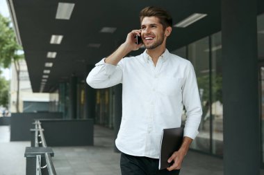 Memnun Avrupalı işadamı cep telefonuyla konuşuyor ve elinde belgeler var. Şehirdeki modern ofis binasının arka planında duran genç adam. Gülümseyen sakallı adam beyaz gömlek ve kot pantolon giyiyor. Gündüz