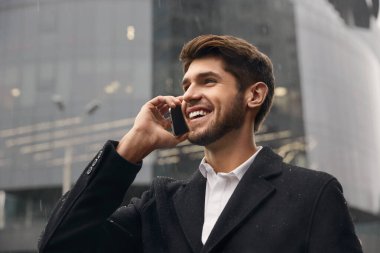 Genç gülümseyen Avrupalı işadamı şehirde cep telefonuyla konuşuyor. Uzaktan kumanda kavramı. Modern başarılı adam. Palto giyen sakallı şık adam. Yağmurlu gün
