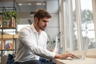 Genç Avrupalı işadamı dizüstü bilgisayarda bir şey izliyor ve elinde belge tutuyor. Modern başarılı adam kavramı. Resmi kıyafetler giyen sakallı şık adam. Ofis içi. Gündüz