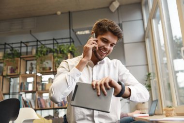 Genç gülümseyen Avrupalı işadamı cep telefonuyla konuşuyor ve kol saatinde zaman geçiriyor. Modern başarılı adam kavramı. Resmi kıyafetler giyen sakallı şık adam. Ofis içi. Gündüz