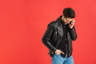 Gülümseyen beyaz sakallı, deri ceketli esmer adam kırmızı arka planda tek başına cep telefonuyla konuşuyor.